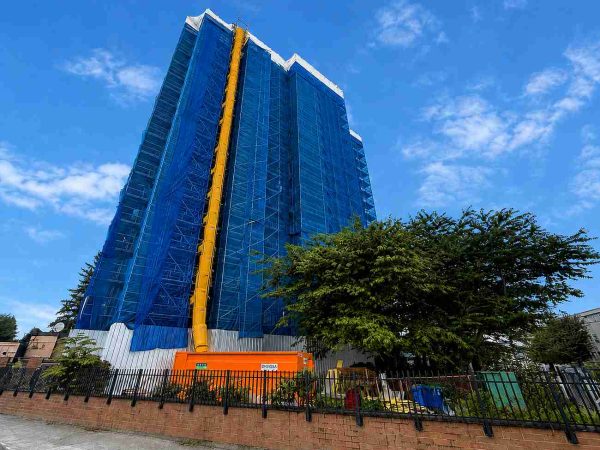 Reliable Scaffolding high-rise building wrapped in blue construction netting with suspended access system and exterior restoration work under a bright blue sky.